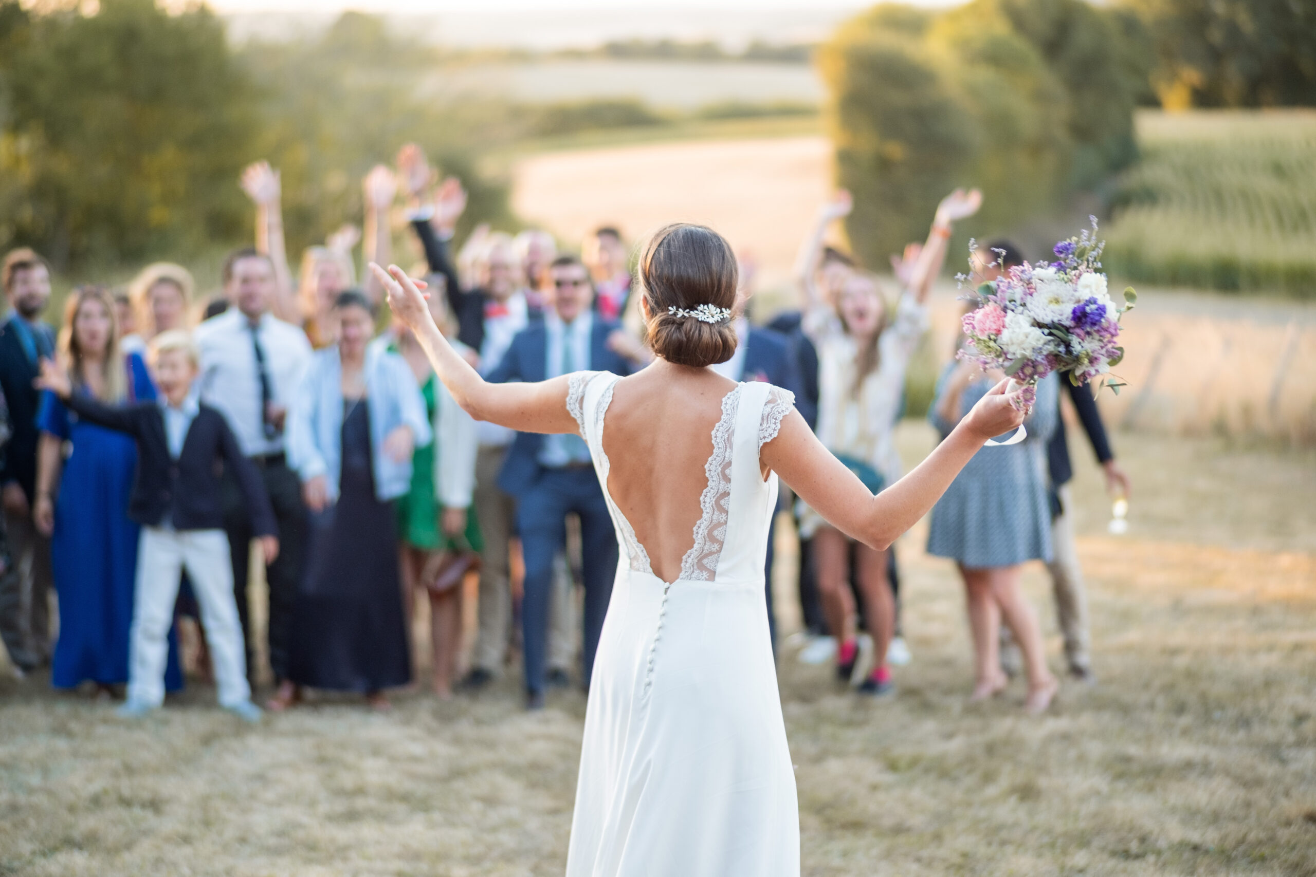 Une mariée de dos qui s'apprête à jeter le bouquet de fleurs qu'elle tient dans sa main droite, avec en fond une foule de gens prêt à le récupérer