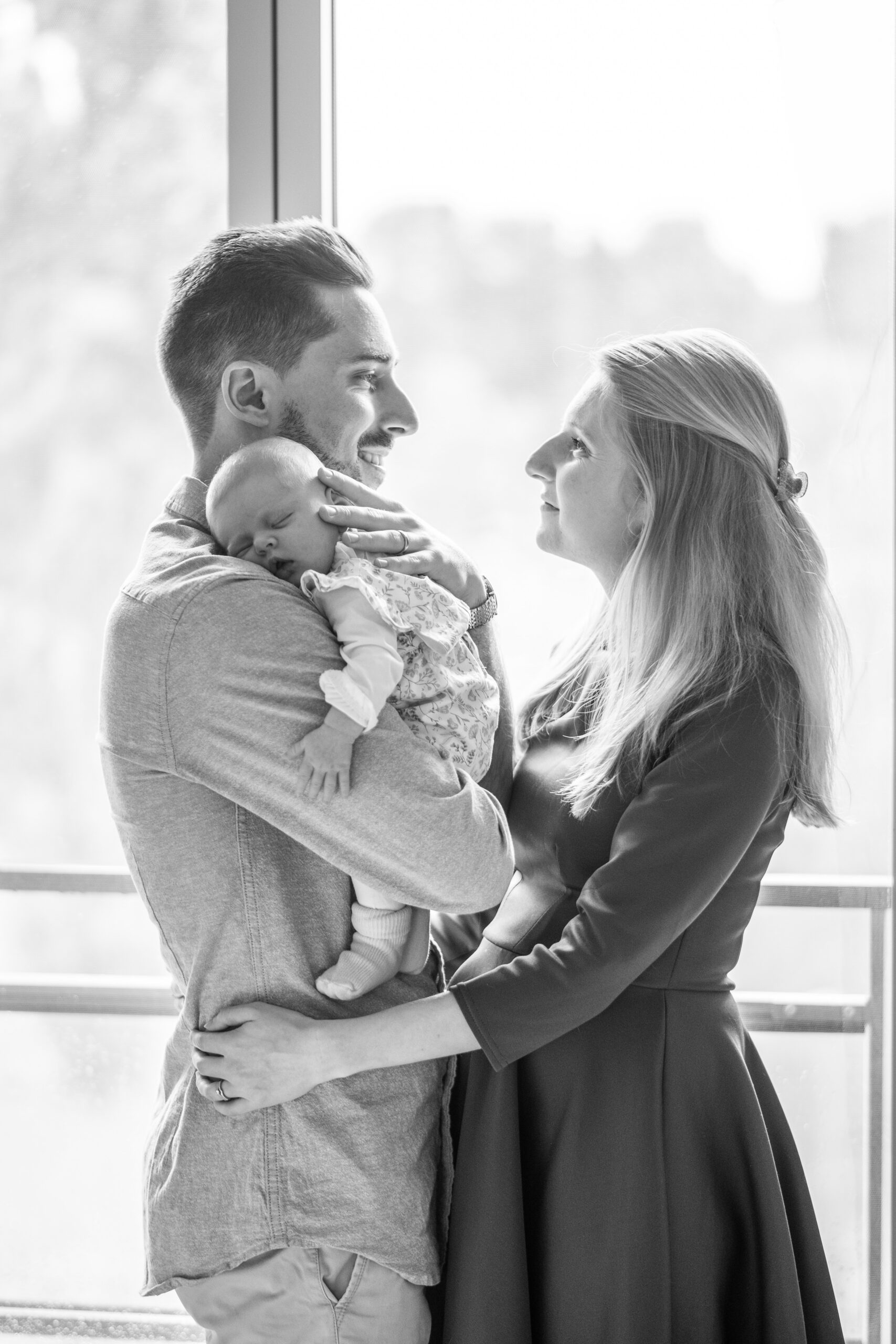 Un homme tient son nouveau né contre lui, et une femme qui se tient devant lui le regarde en souriant, la photo est en noir et blanc